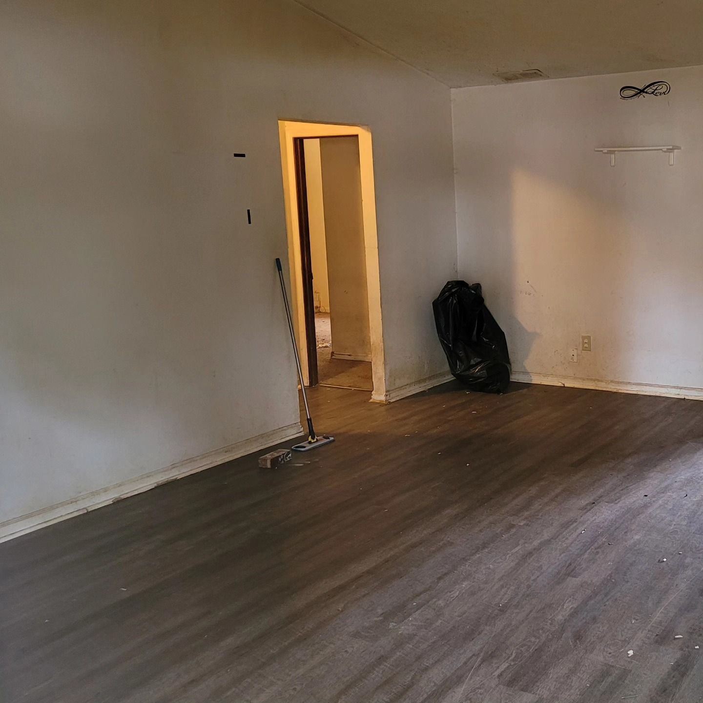Empty room with wooden floors, open doorway, black trash bag, and exposed drywall.