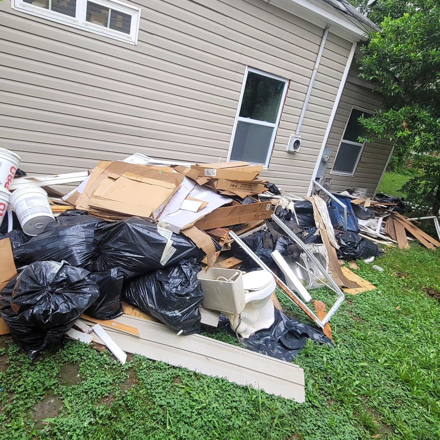 Pile of trash bags, cardboard boxes, and debris beside a beige house with windows, on a green lawn.