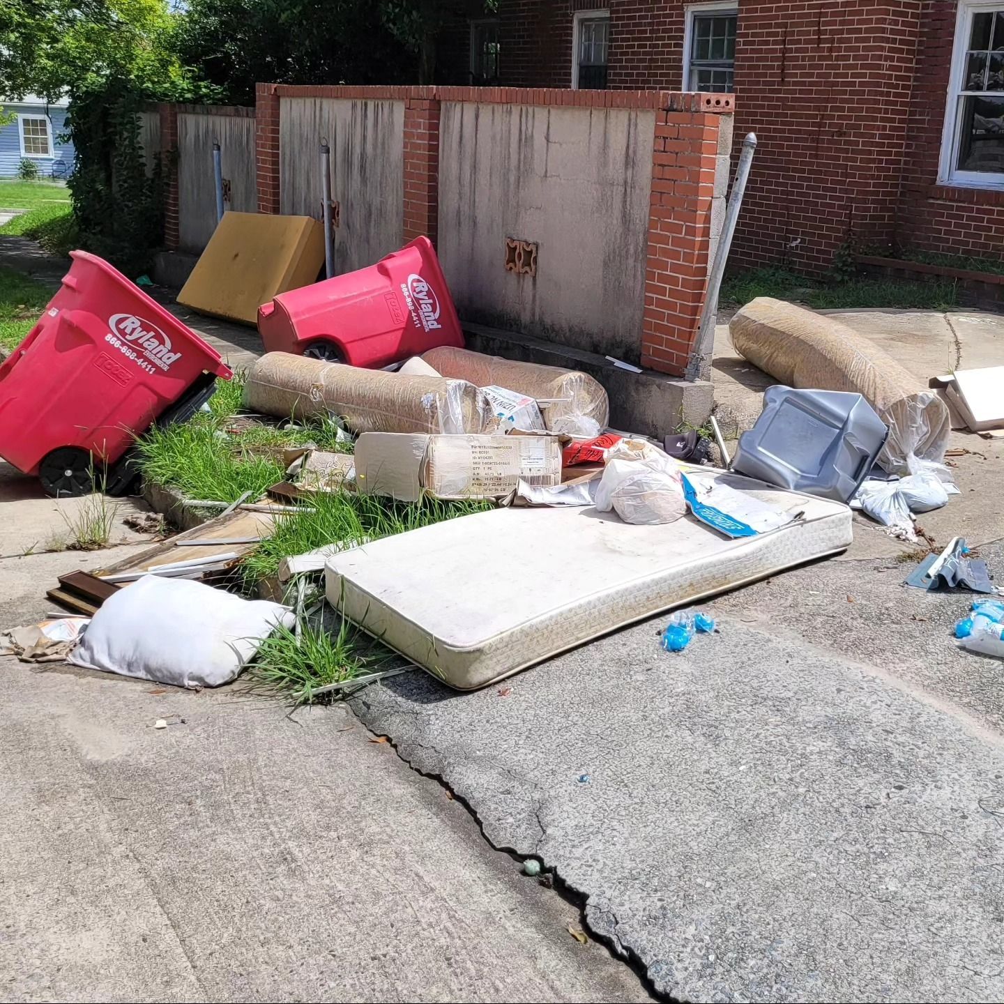 A pile of discarded trash including a mattress, trash bins, and debris, on a sidewalk next to a brick building.