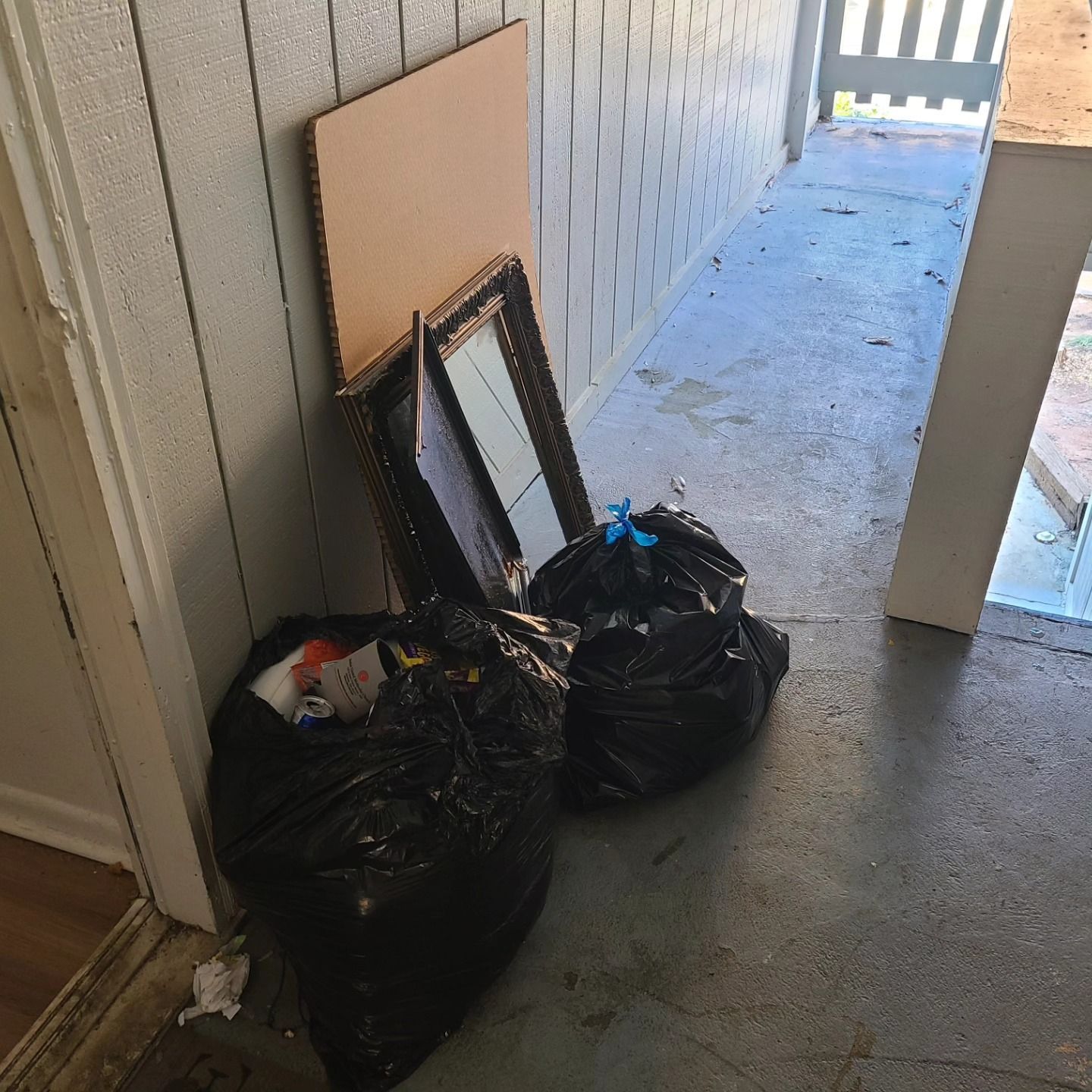 Two black trash bags, cardboard, and a framed mirror lean against a wall on a porch.