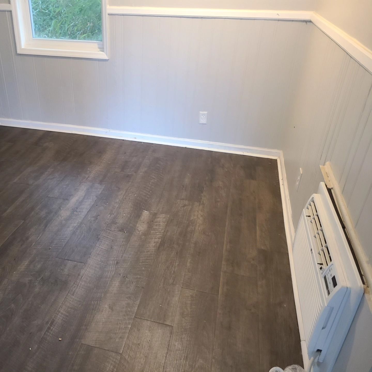Room with grey flooring and walls, small window, white trim and an air conditioner unit.