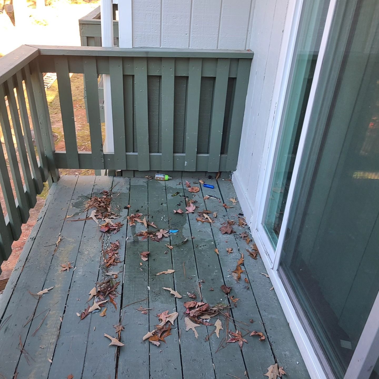 Dirty wooden balcony with leaves, next to a sliding glass door and green painted railing.