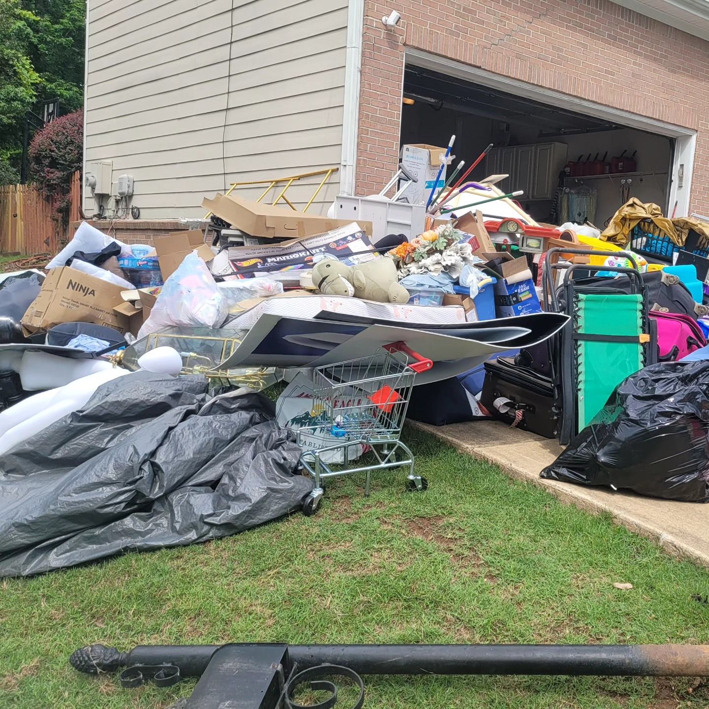 Pile of discarded items next to a garage, including boxes, trash bags, and a shopping cart, on grass.