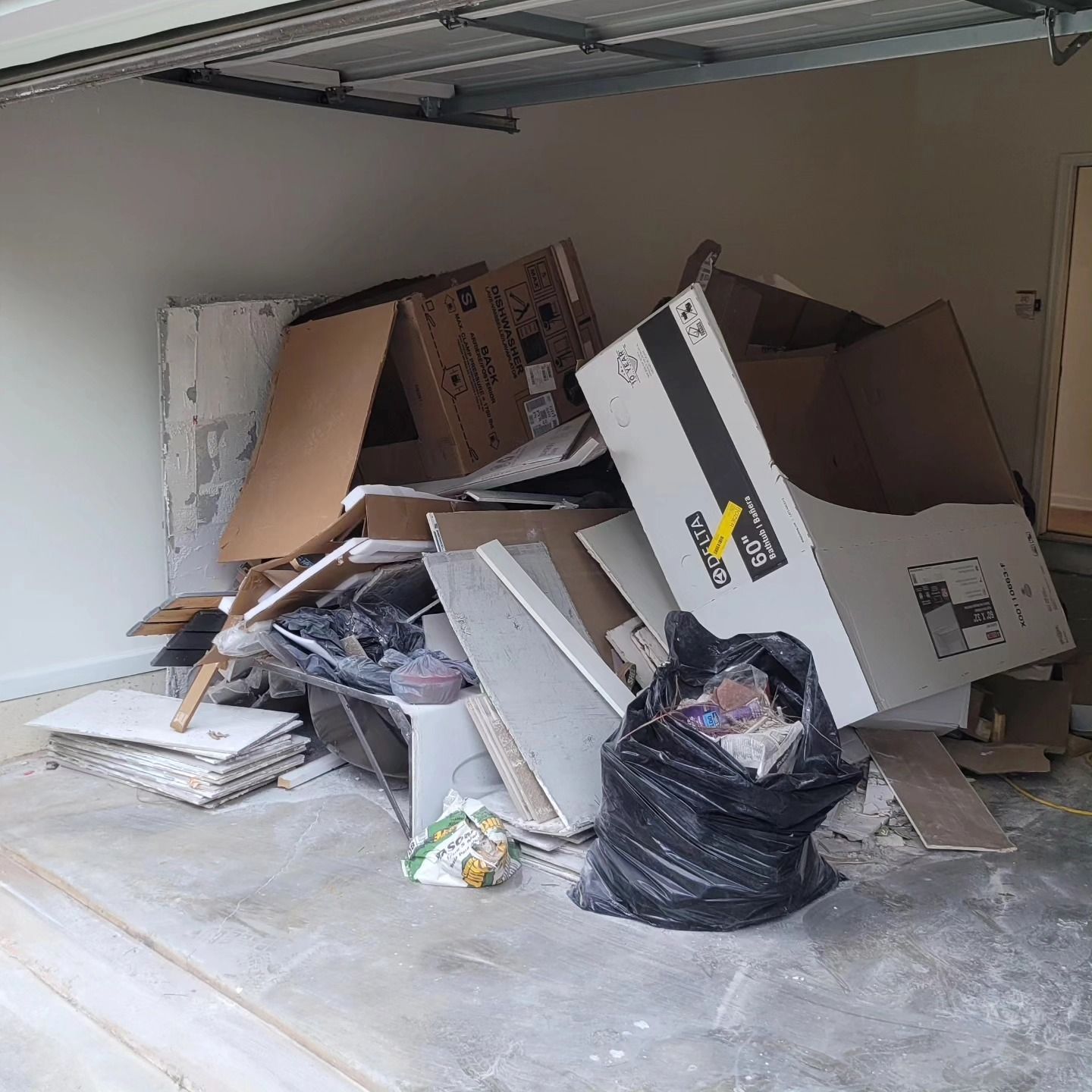 Pile of construction debris in a garage, including cardboard boxes, drywall, and a black trash bag.