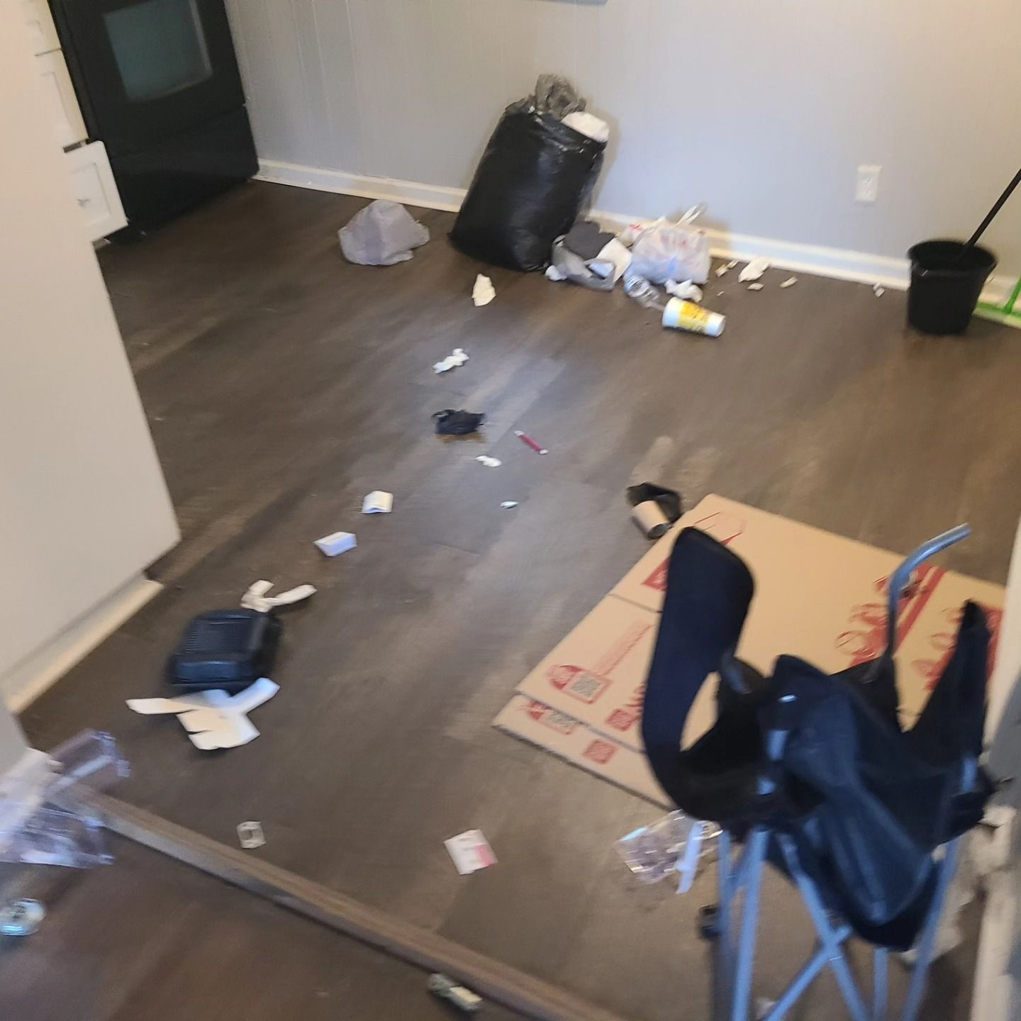 A room with a scattered trash. Dark floors, black trash bag, discarded items. Black chair and a box.