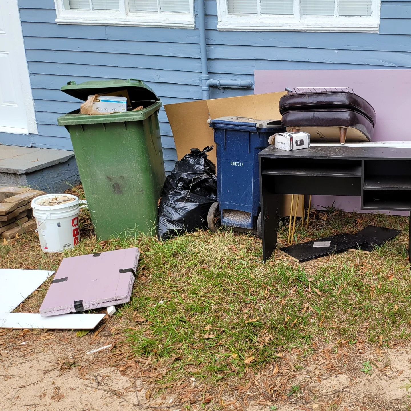 Exterior view with trash cans, discarded furniture, and debris on a grassy area next to a blue house.