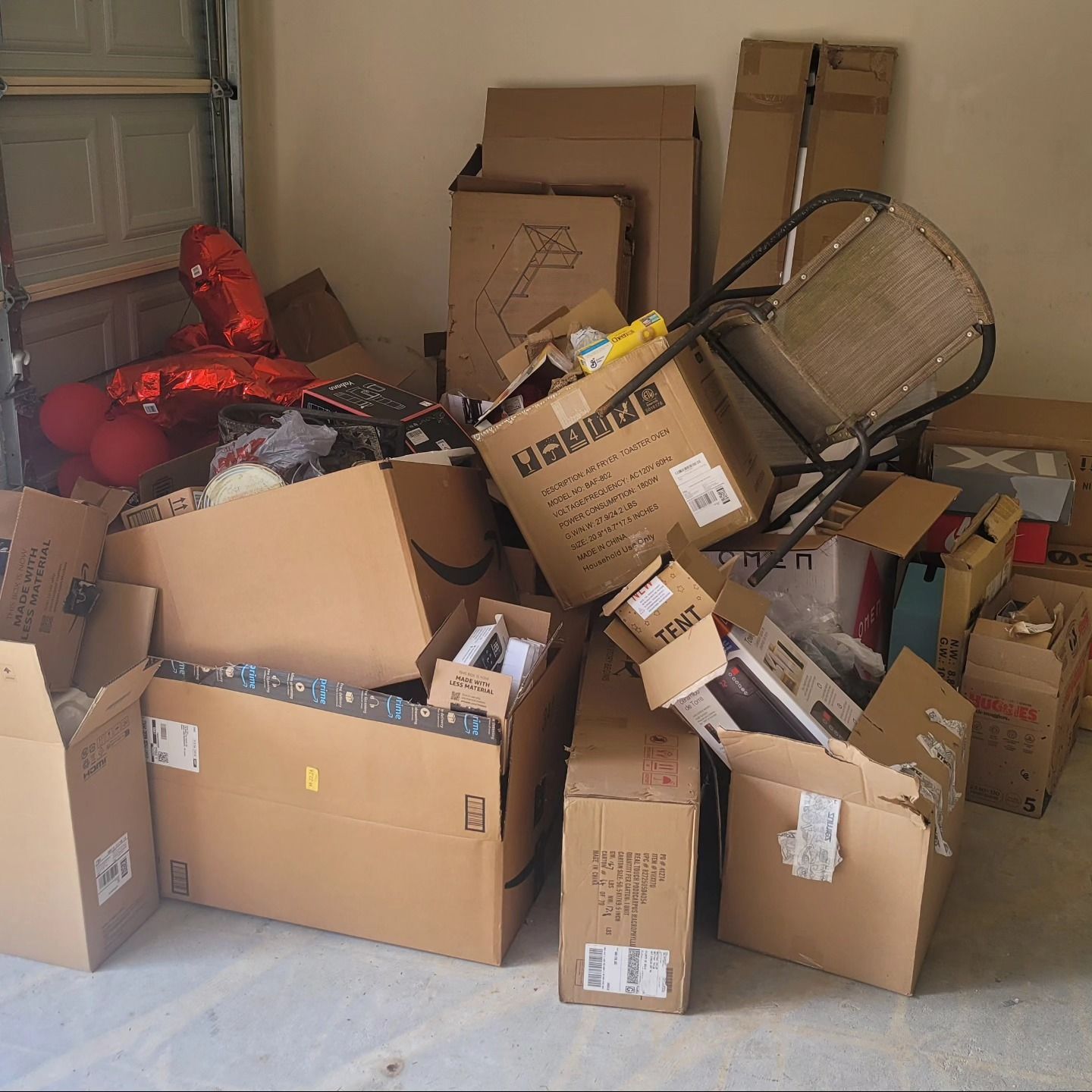 Pile of cardboard boxes and various items inside a garage, including a chair and red balloon decoration.