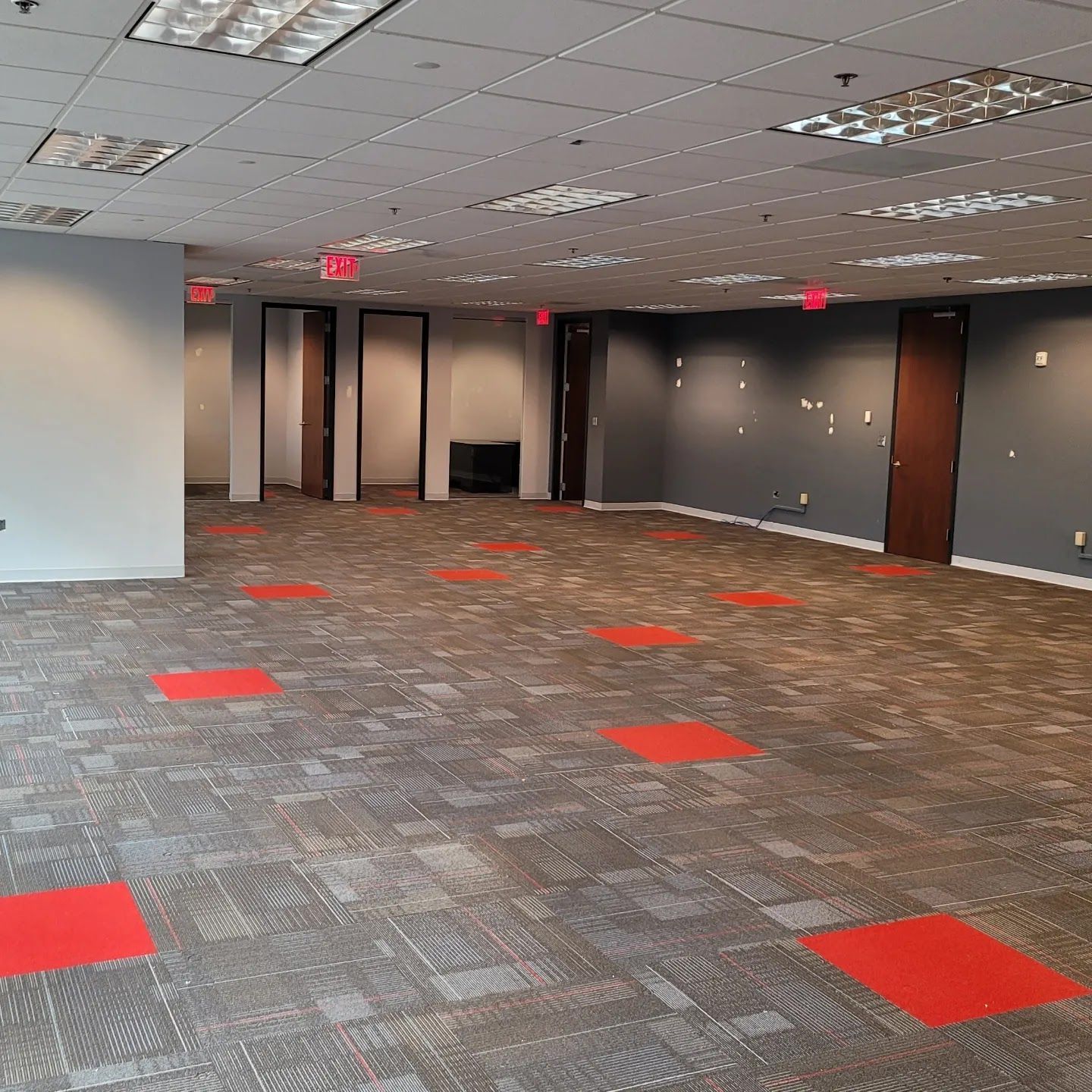 Empty office space with gray carpet, red squares, and several closed doors. Fluorescent lights on the ceiling.