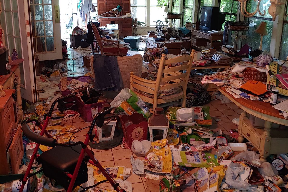 Cluttered living room: debris and items cover the floor and furniture, walker in foreground, windows and door at back.
