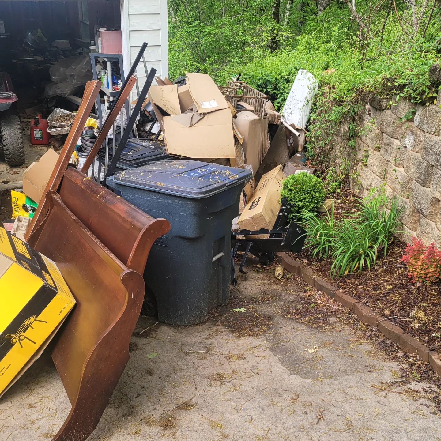 Pile of trash including furniture and cardboard boxes next to a blue trash can in a yard.