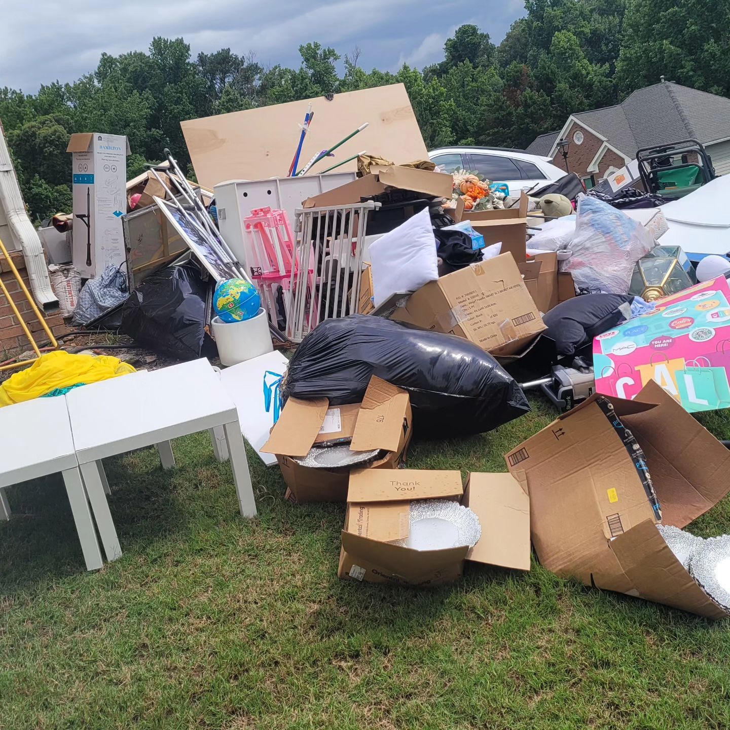 Pile of discarded household items, boxes, furniture, and trash on a lawn, next to a home.