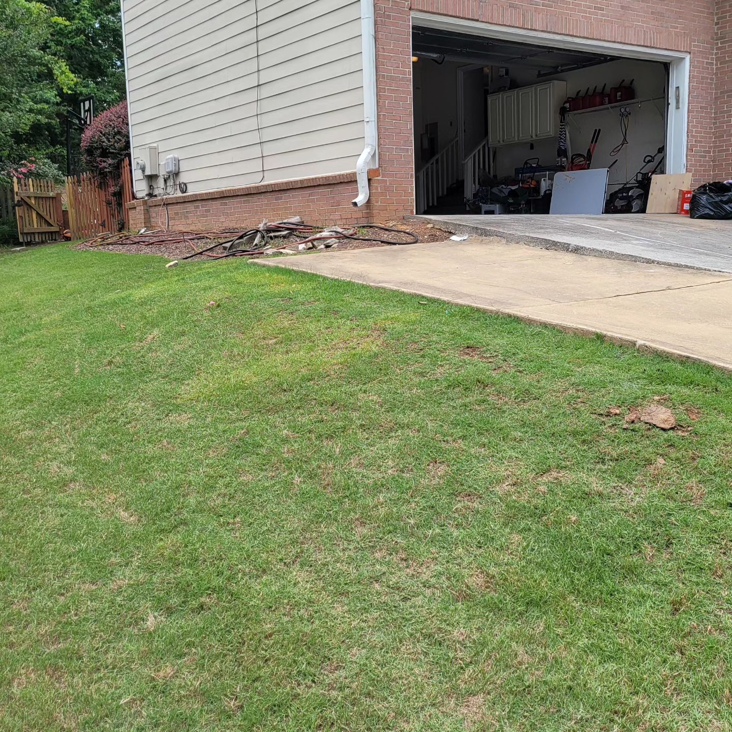 Green lawn slopes towards a driveway and garage. Beige house with brick base and open garage.