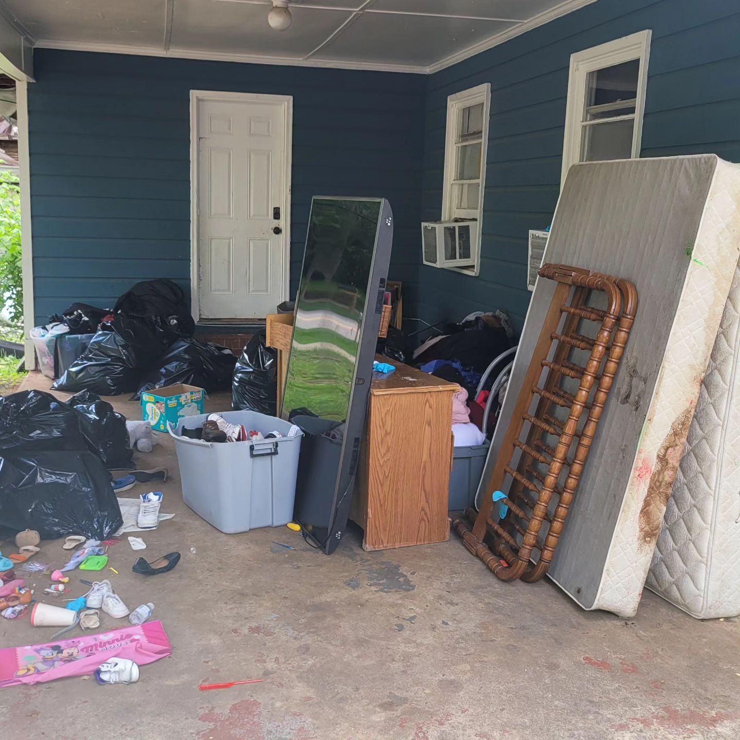 Garage with trash bags, mattress, furniture, and a door against a blue house.
