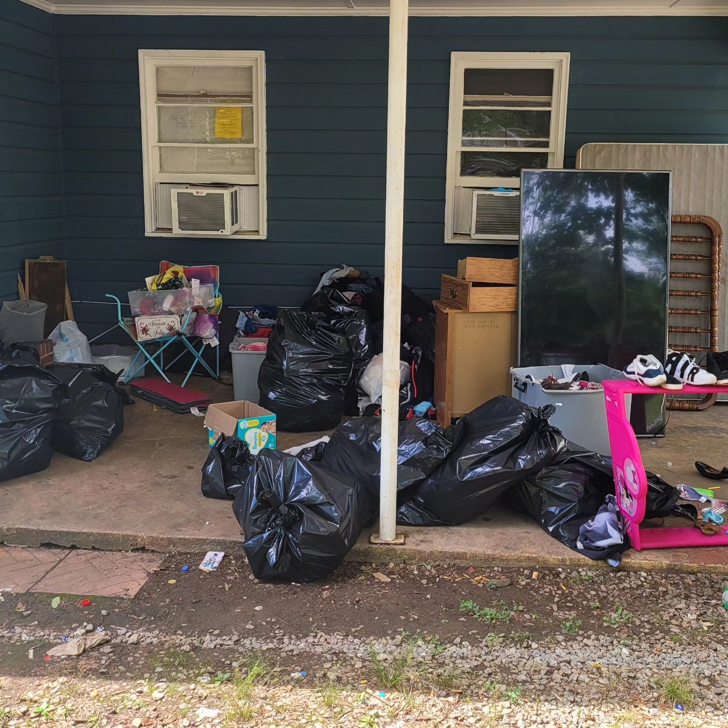 Cluttered porch with black trash bags, cardboard boxes, and miscellaneous items; two windows with air conditioners.