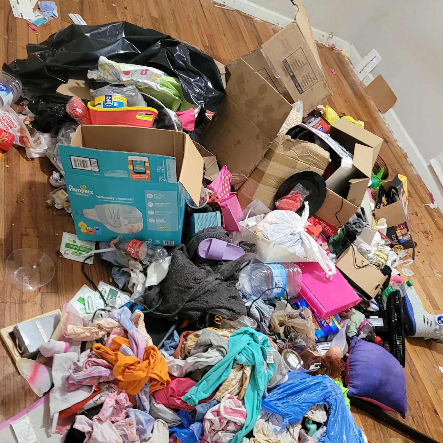 Pile of garbage, including boxes, trash bags, and clothes, strewn on a wooden floor.