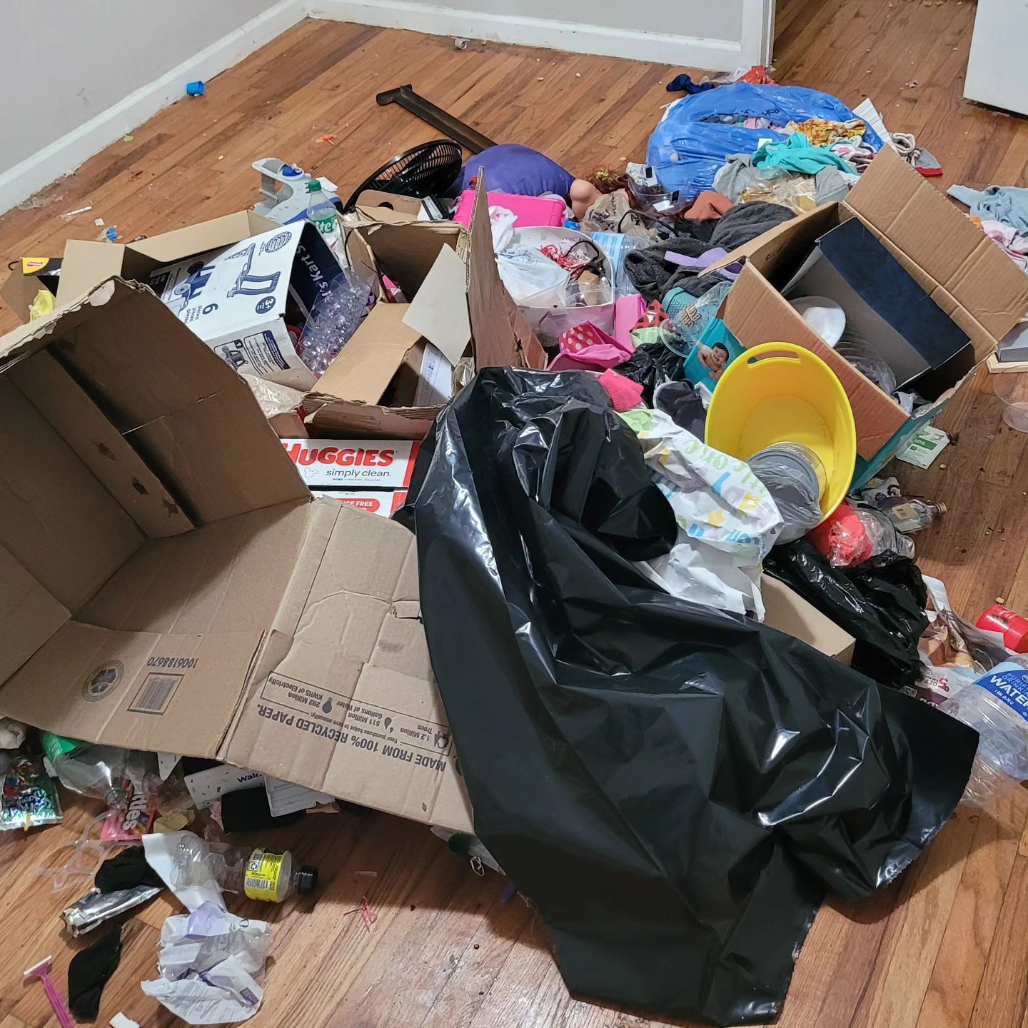 Room in disarray: cardboard boxes, clothes, trash bag, and various items strewn on hardwood floor.