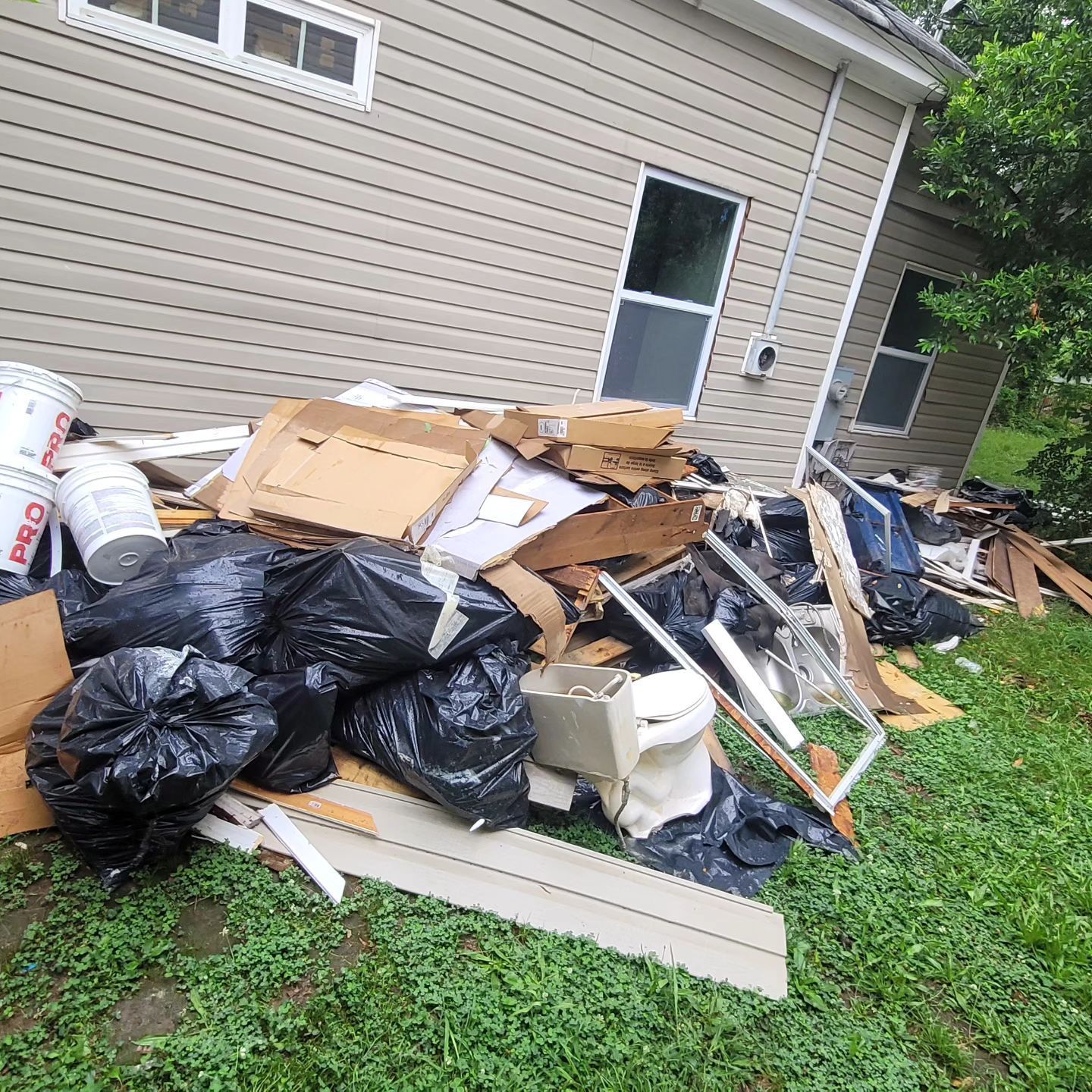 Pile of trash and debris in front of a house, including black bags, cardboard, and construction materials.