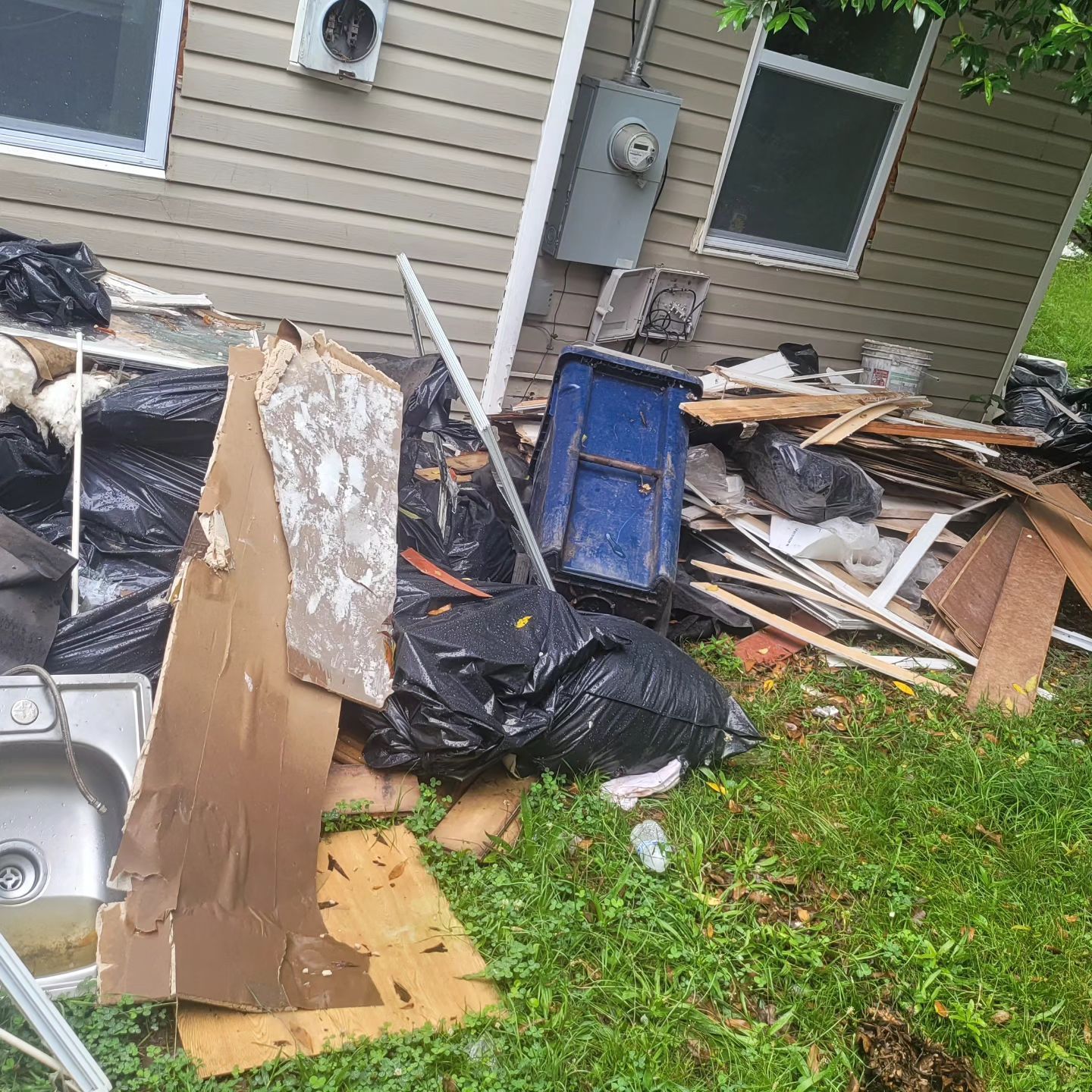 Pile of trash, including wood and black bags, next to a house with an air conditioner.