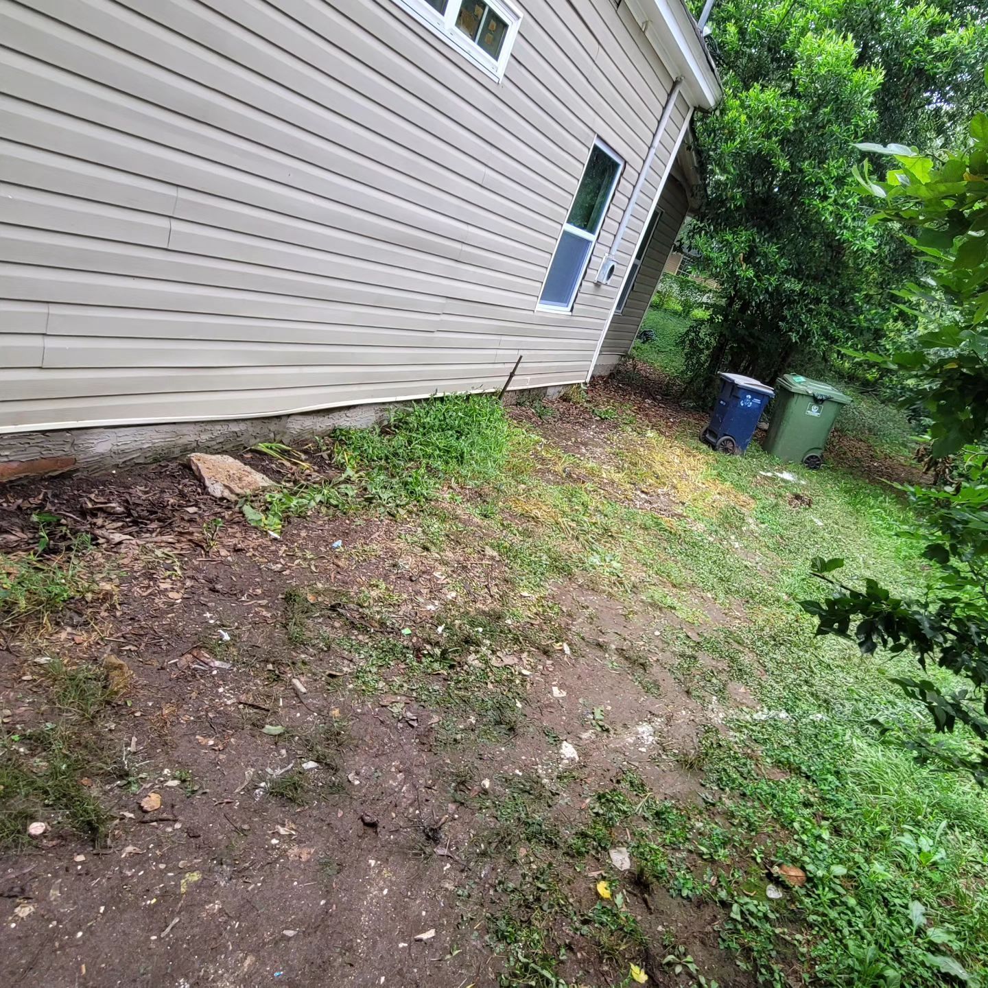 Side of a house with beige siding, dirt, and some grass. Trash cans are in the background.