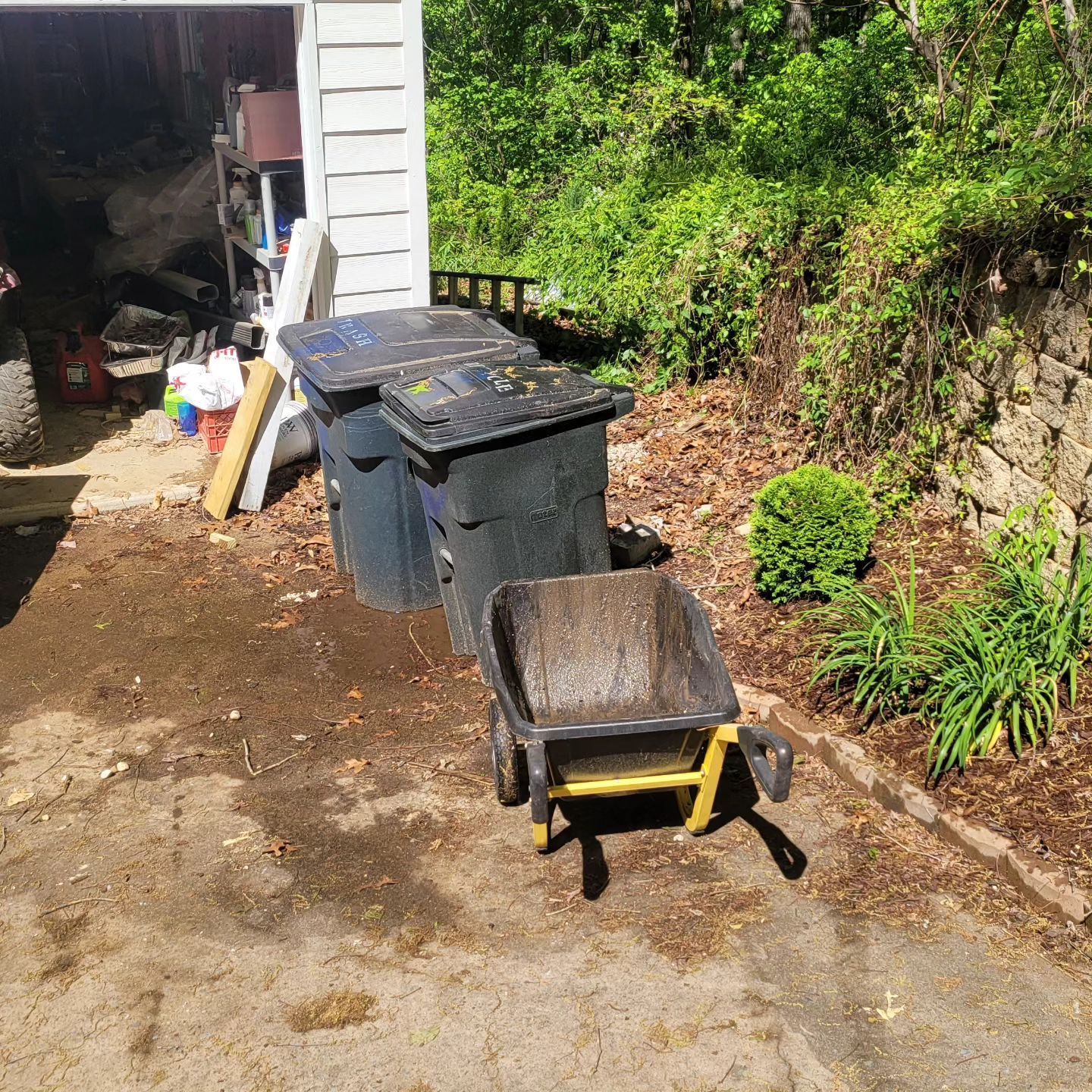 Wheelbarrow and trash cans in a driveway next to a stone wall and wooded area.