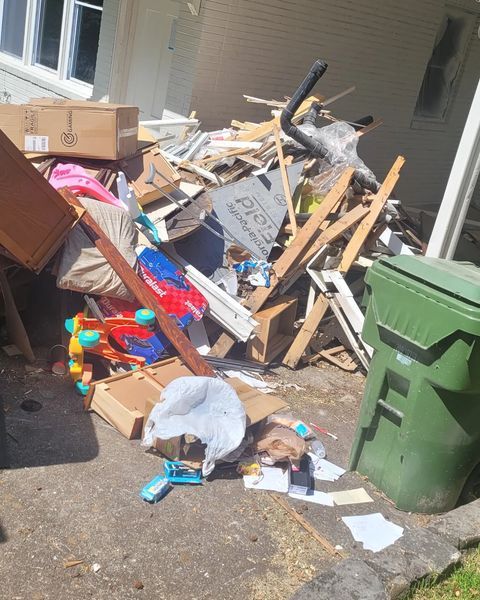Pile of construction debris and trash next to a green trash bin on a concrete surface, near a house.
