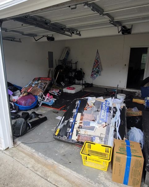 Cluttered garage interior. Various items strewn about, including boxes, baskets, and a large object covered in paper scraps.