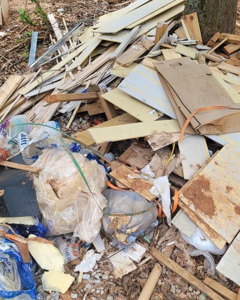 Pile of construction debris and trash in a wooded area, including wood, plastic, and foam.