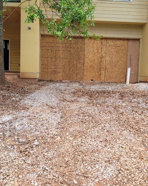 Building with boarded-up garage door. Ground covered in wood chips and debris.
