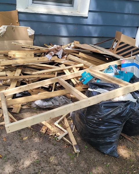 Pile of wooden debris, including pallets and framing, near a blue house wall with a window.