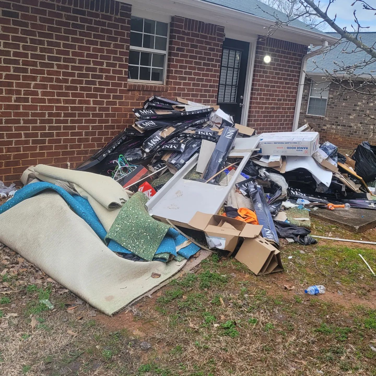 Pile of discarded items next to a brick building. Carpeting, cardboard boxes, and trash overflow on the grass.