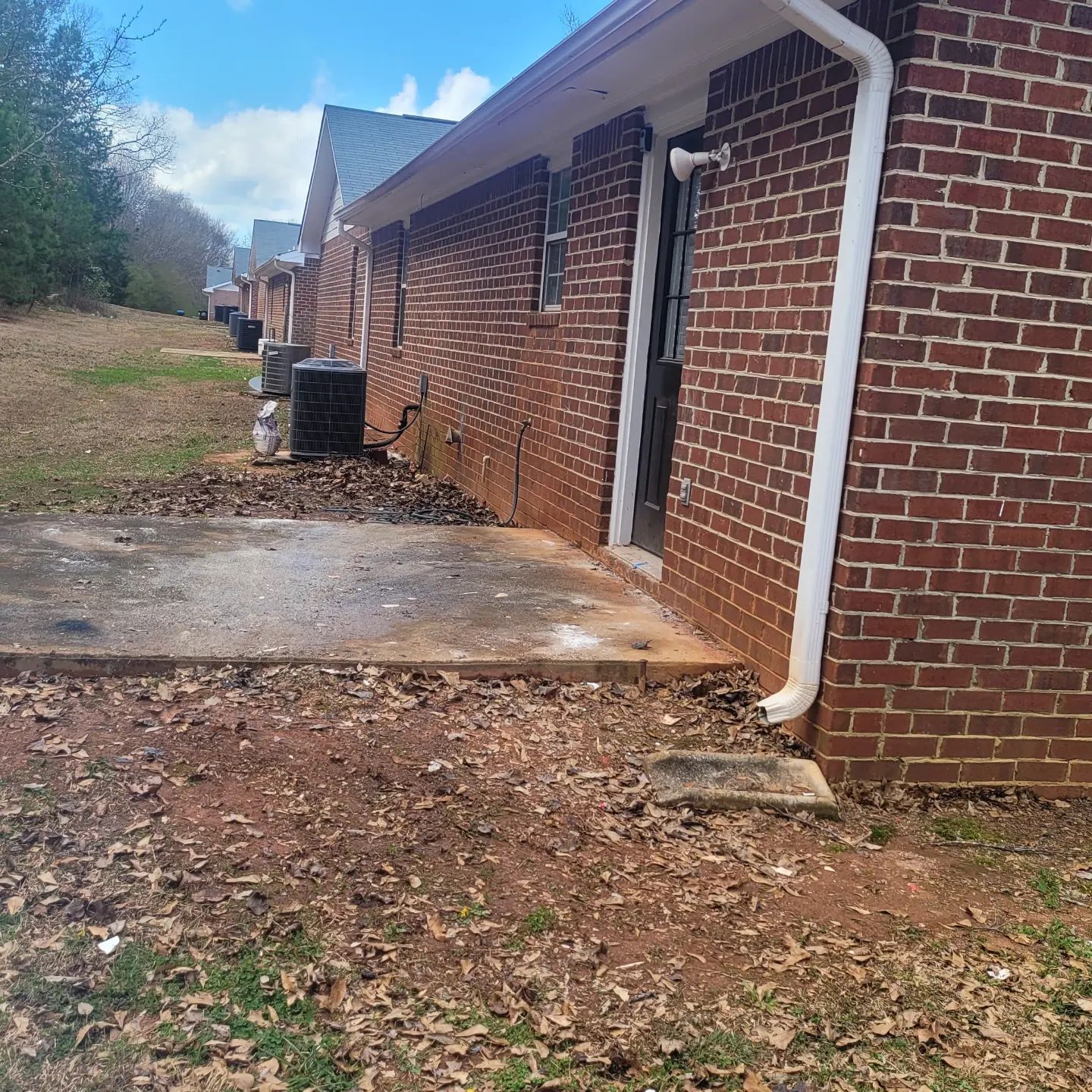 A brick building with a concrete patio covered in leaves. A/C units sit beside the building.