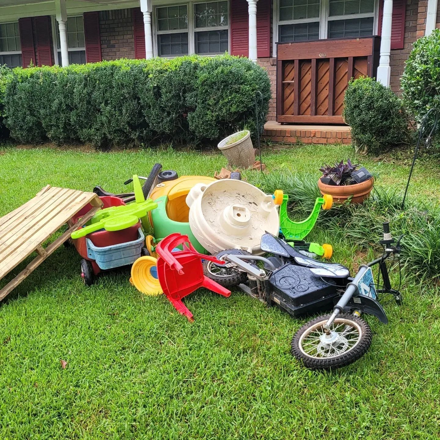 Pile of discarded toys and a pallet on a grassy lawn in front of a house.