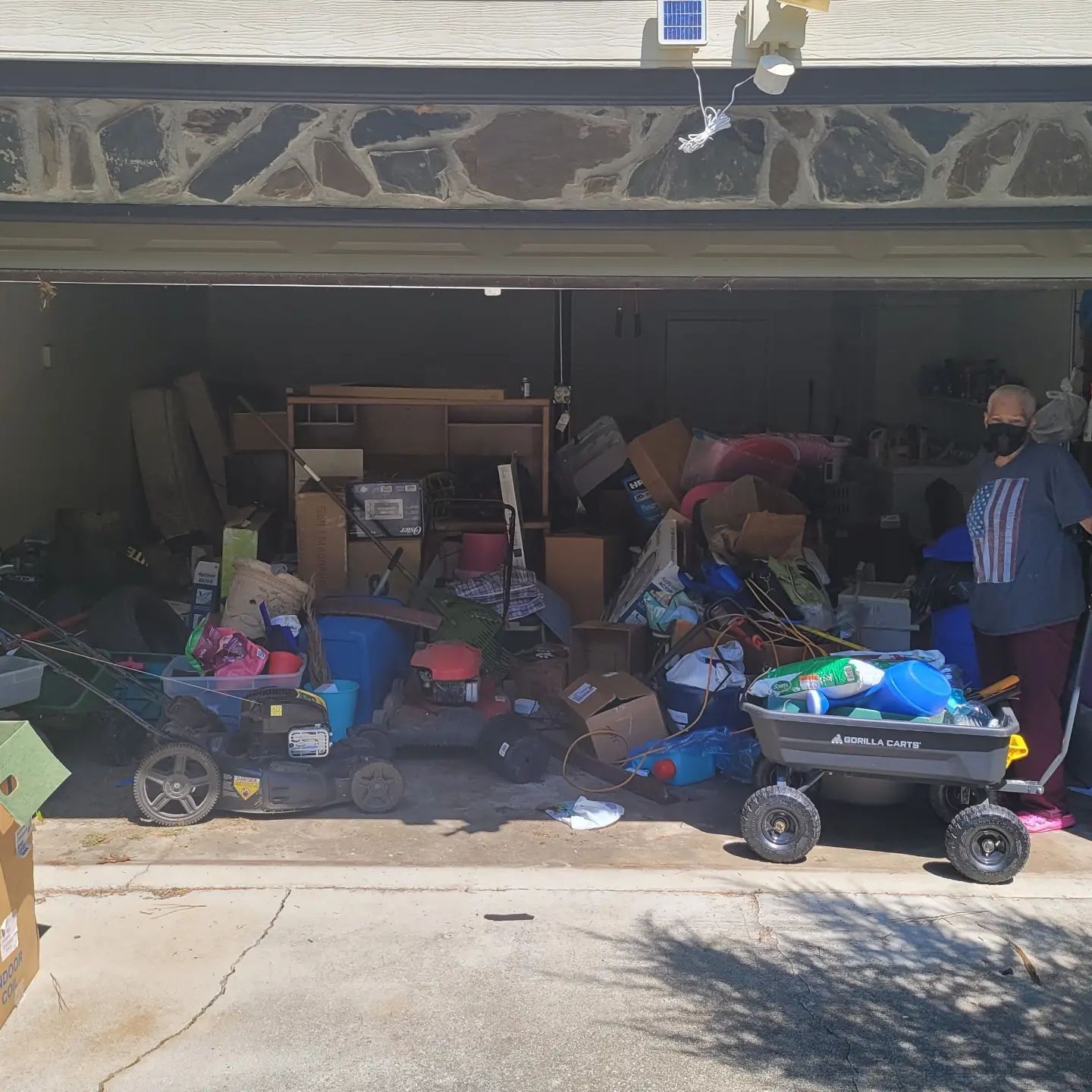 Garage overflowing with items, a person wearing a mask stands on the right. Two wagons and lawn mowers are visible.