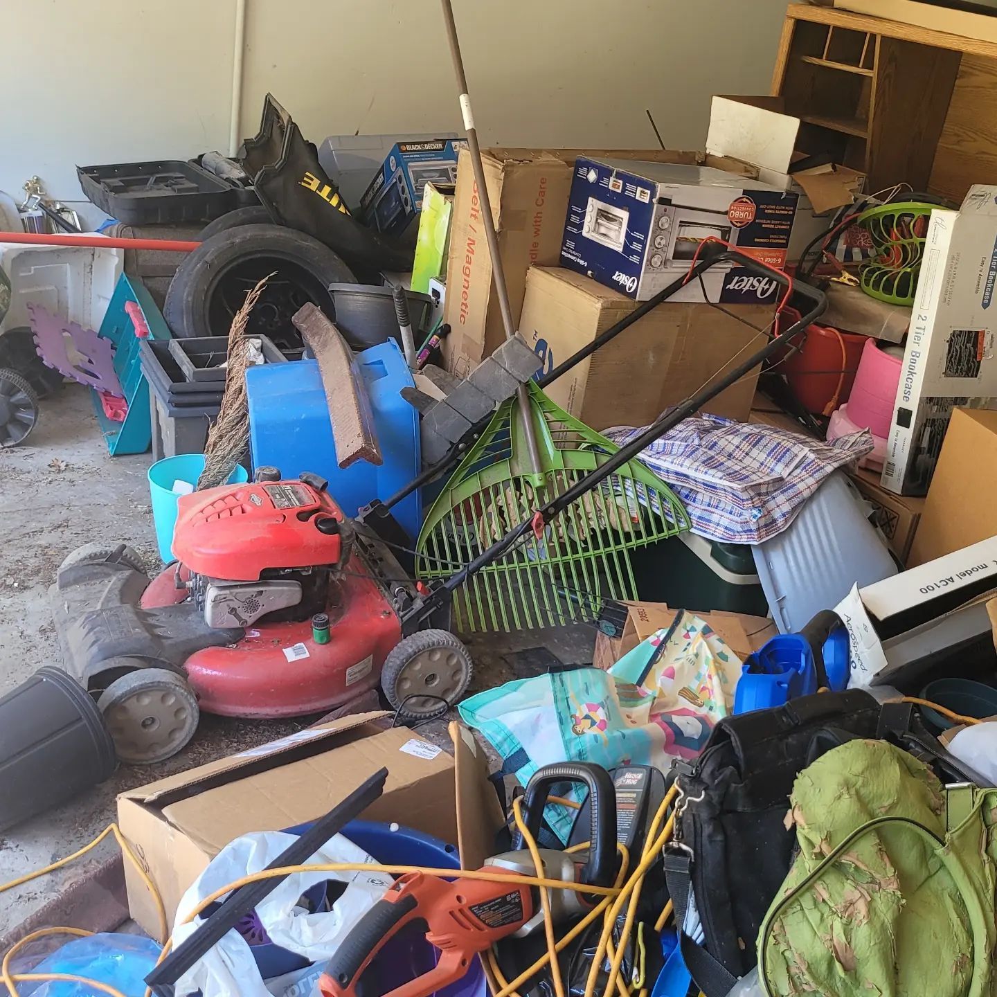 A cluttered garage with a lawnmower, rake, boxes, and various items piled together.