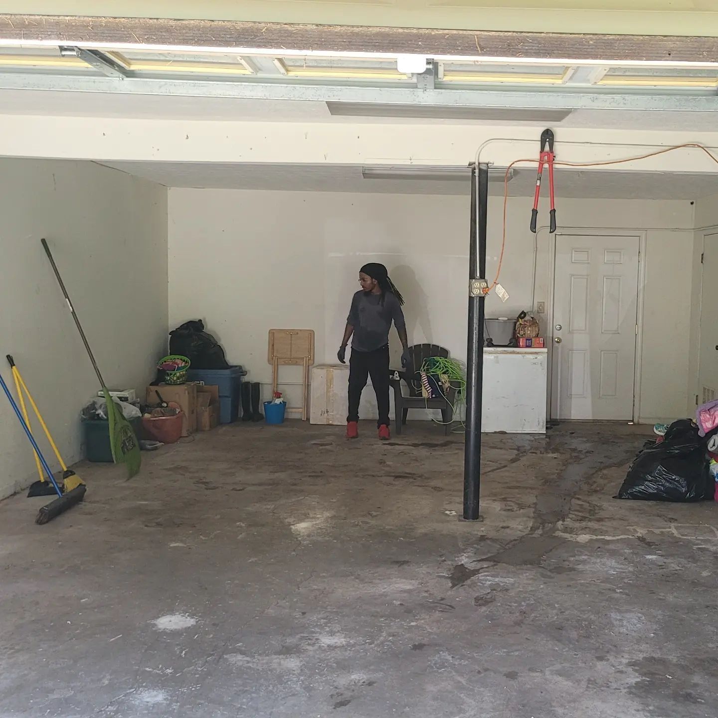 Person standing in a cluttered garage, looking towards the right, with cleaning supplies and bags along the walls.