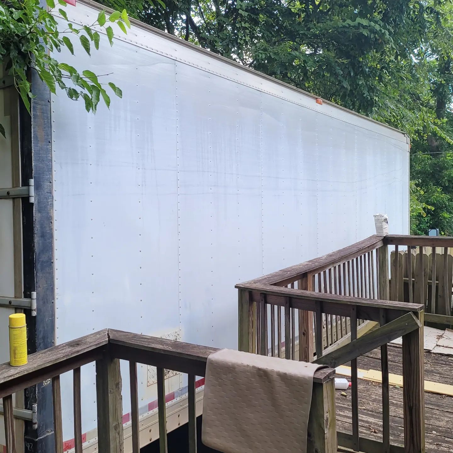 White trailer side beside a wooden deck, trees in the background. A rolled towel on the deck railing.