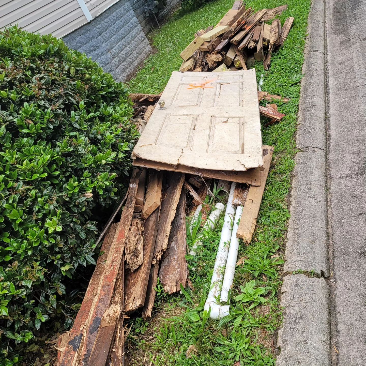 Pile of discarded wood, door, and debris on grass next to a sidewalk and a building.