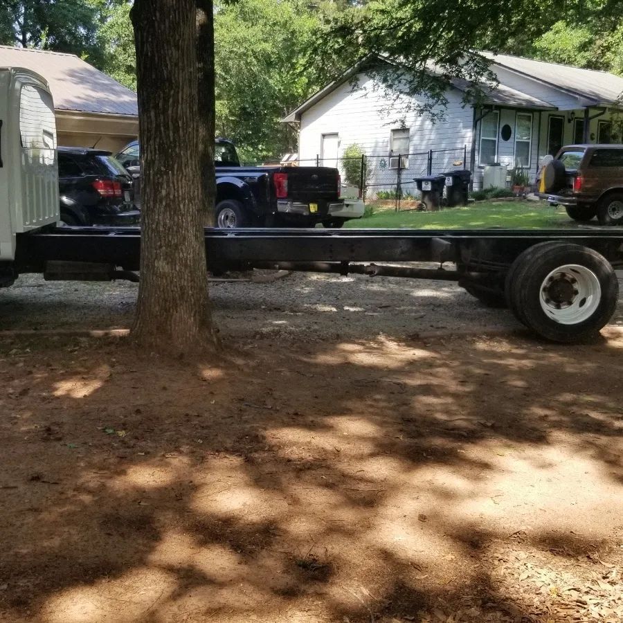 Truck chassis with a tree in front, parked on gravel in a residential area.