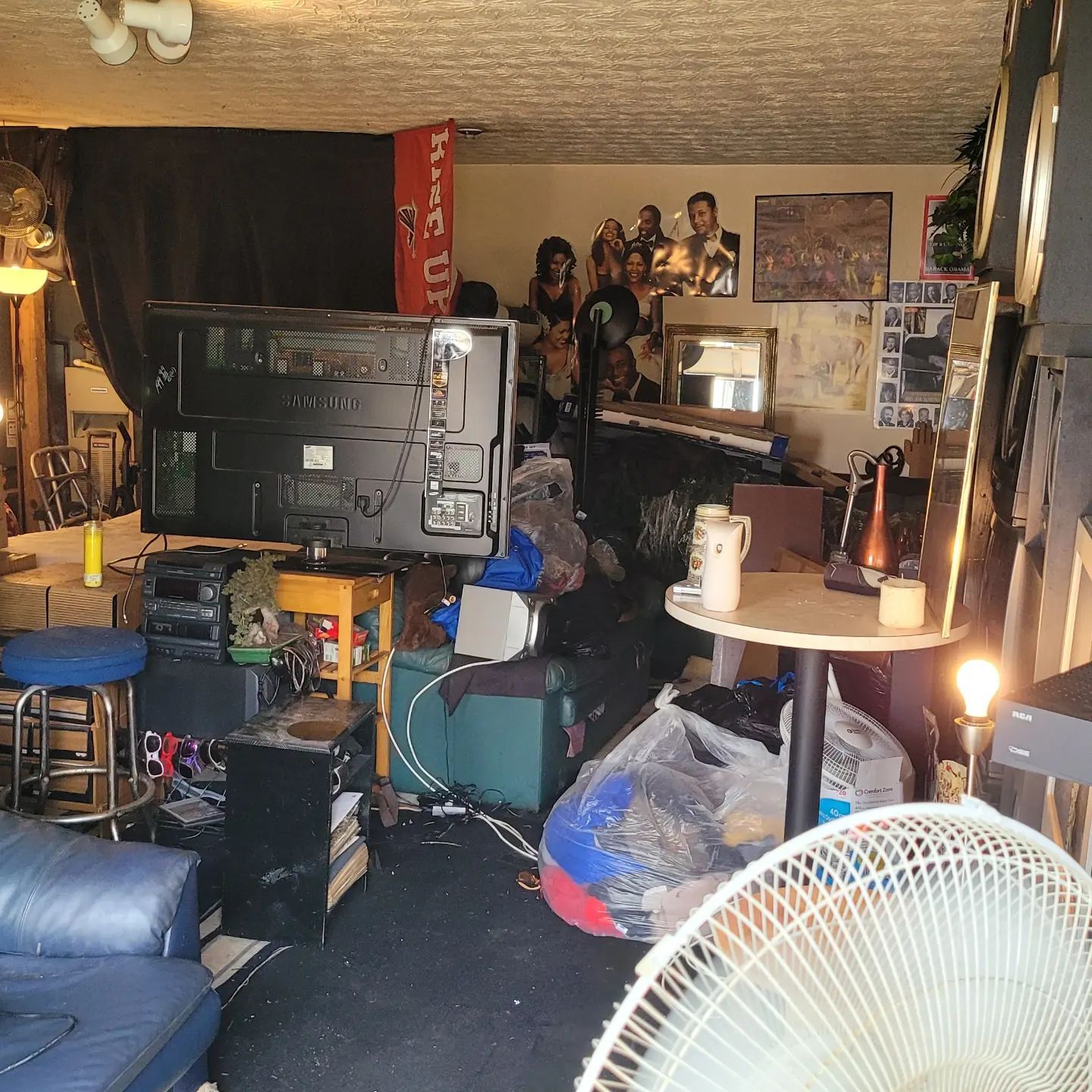 Cluttered living room: TV, furniture, and various objects. A fan is in the foreground.