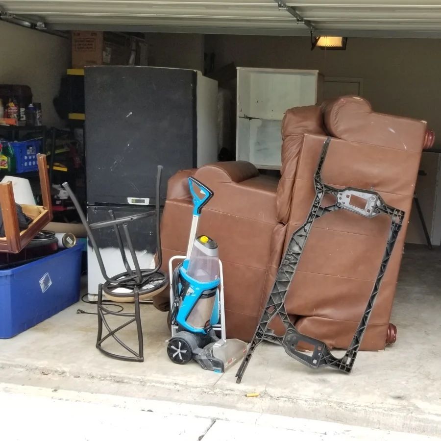 Garage filled with discarded items: a brown chair, vacuum, appliances, and broken furniture.