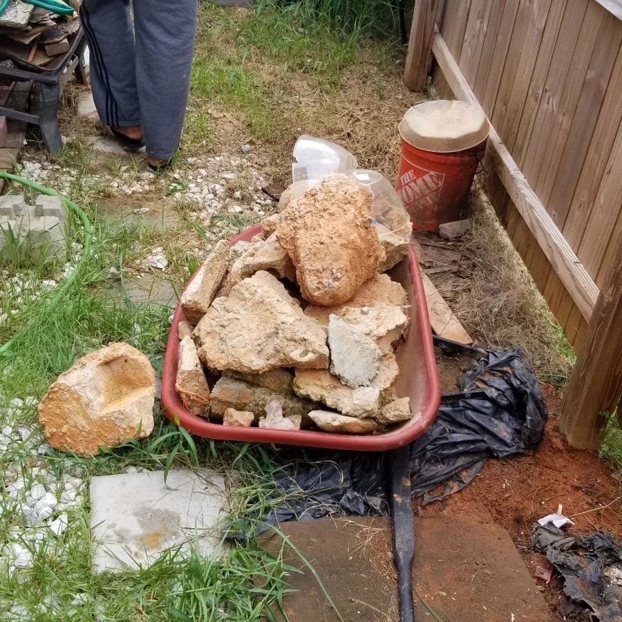 Red wheelbarrow filled with broken concrete and rocks next to wooden fence. Person standing nearby.
