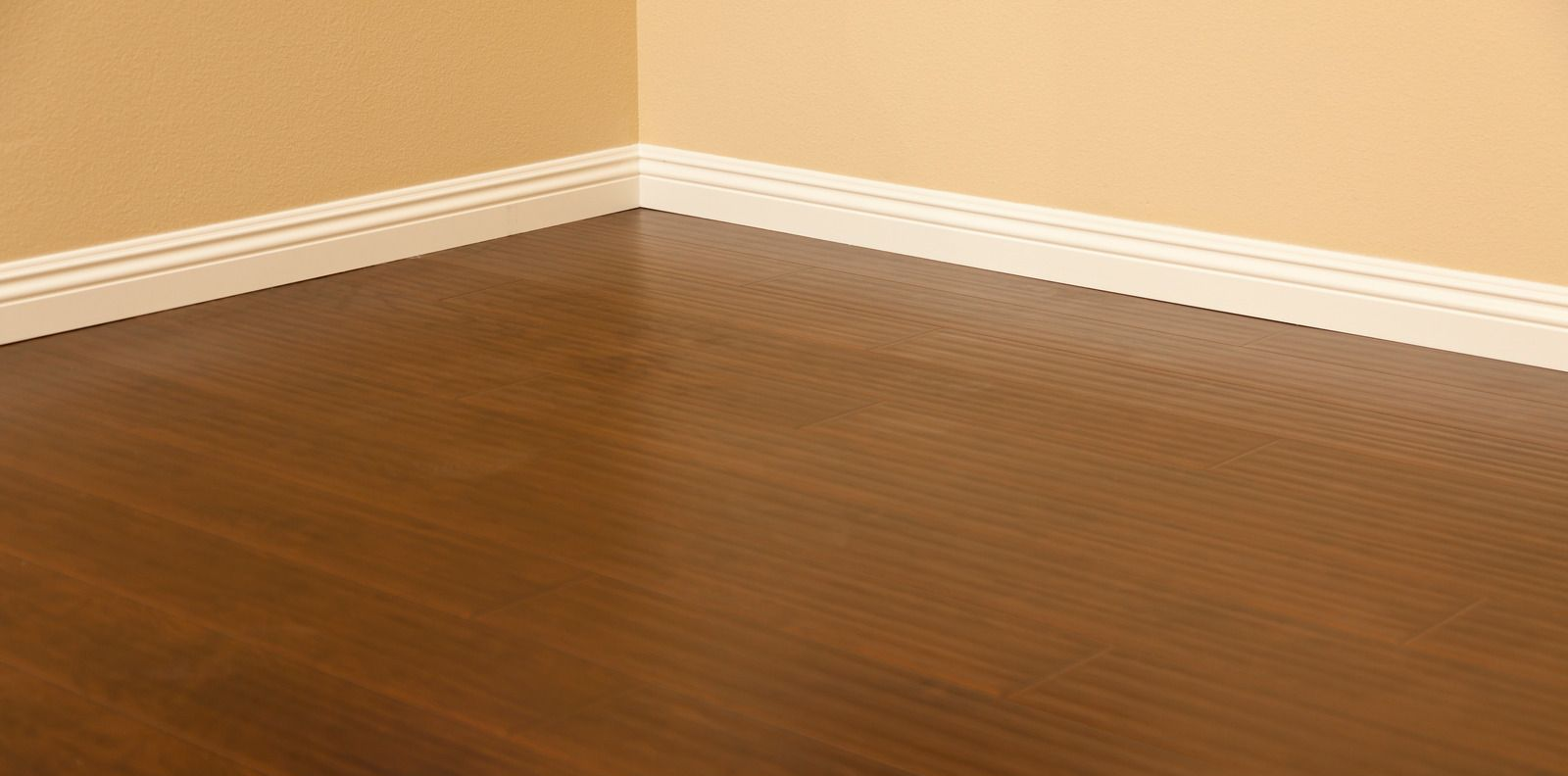 Corner of a room with brown wood flooring, off-white baseboards, and tan walls.