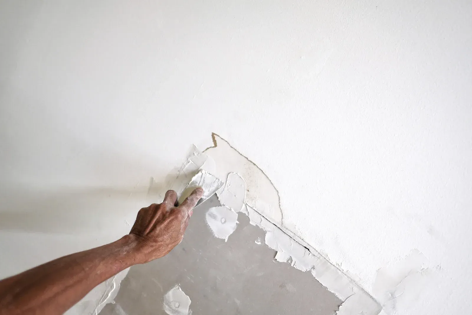 Hand using a putty knife to apply plaster to a white wall.