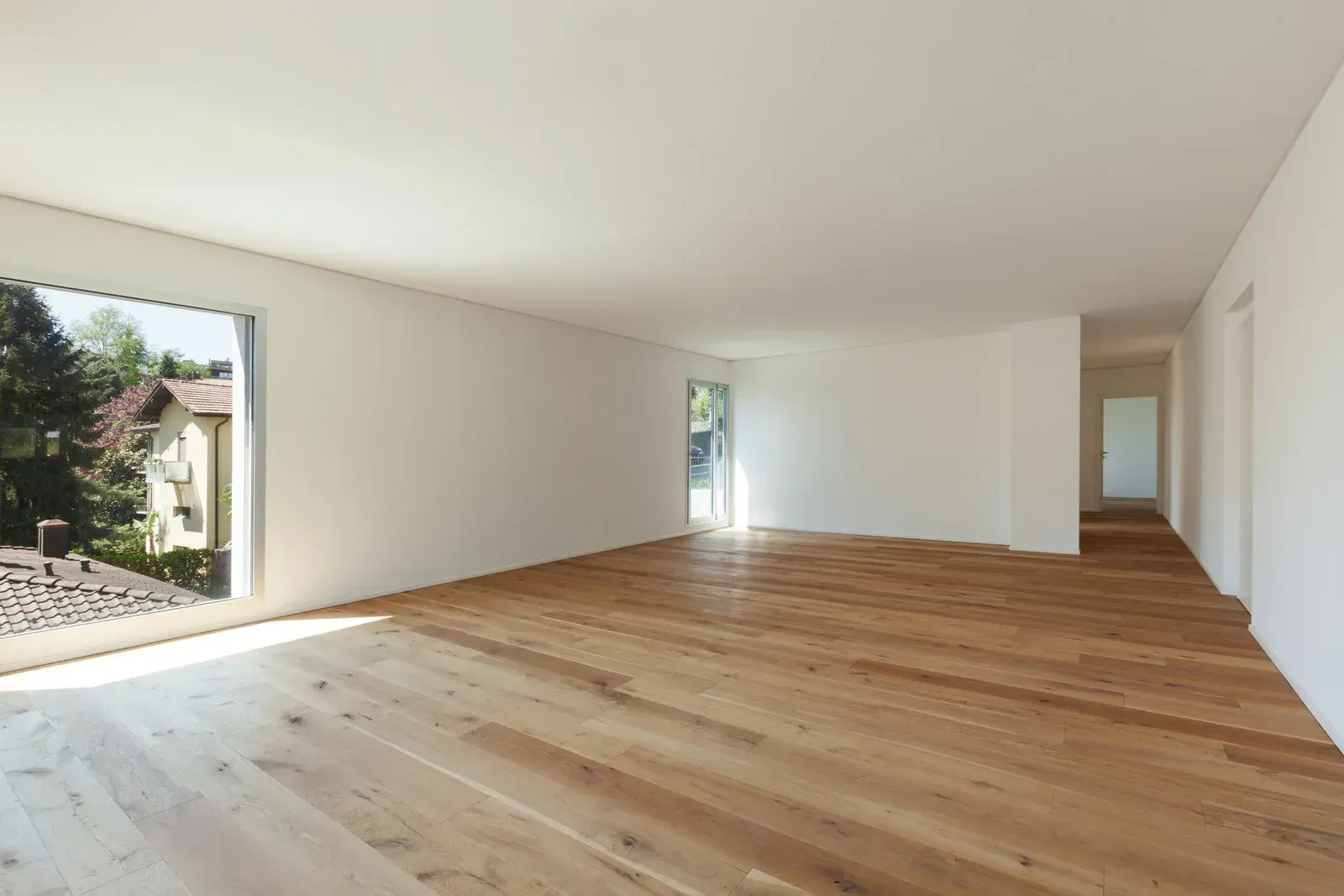 Empty room with hardwood floors, white walls, and a large window with a view of greenery.