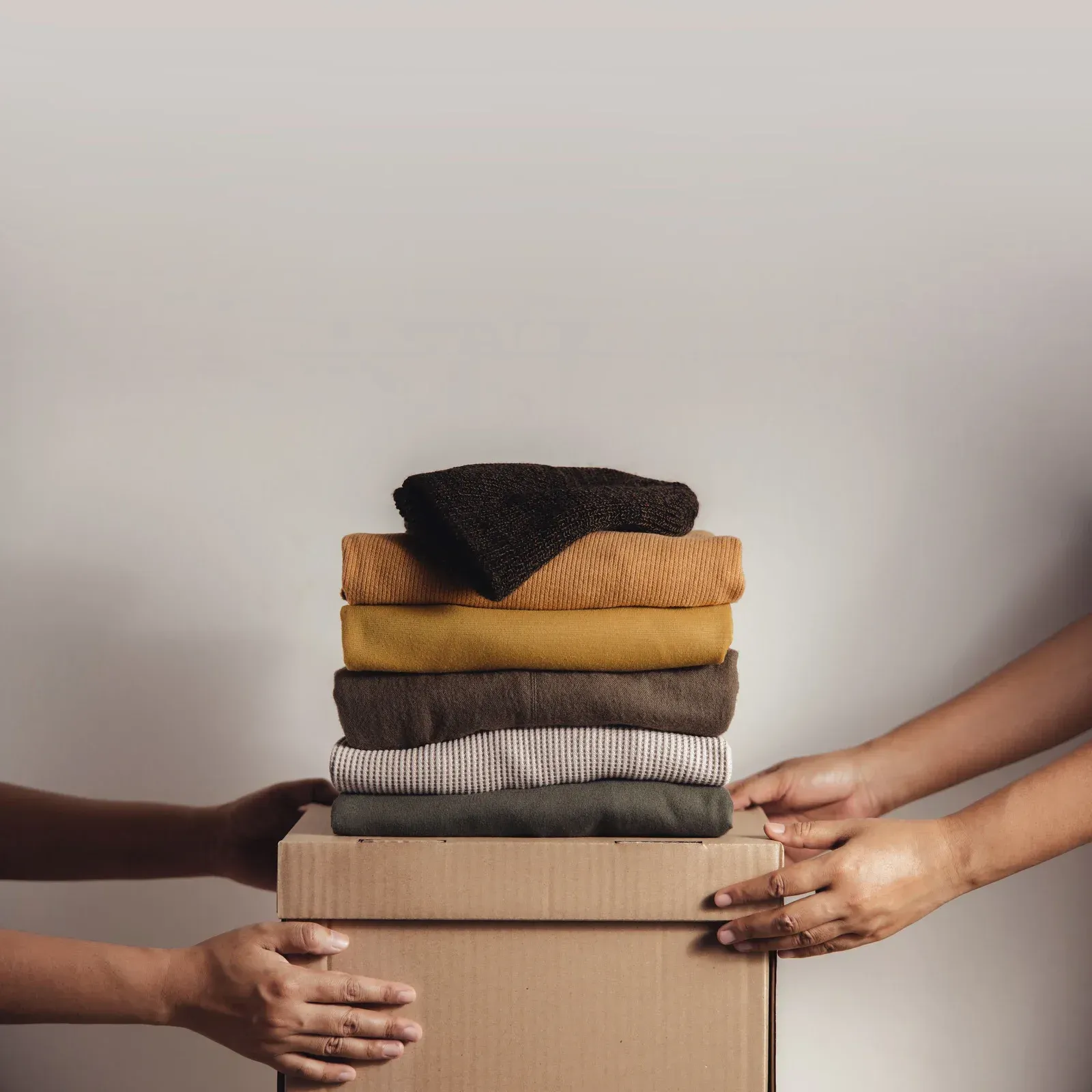 Hands holding a cardboard box with a stack of folded clothes on top. Against a white background.