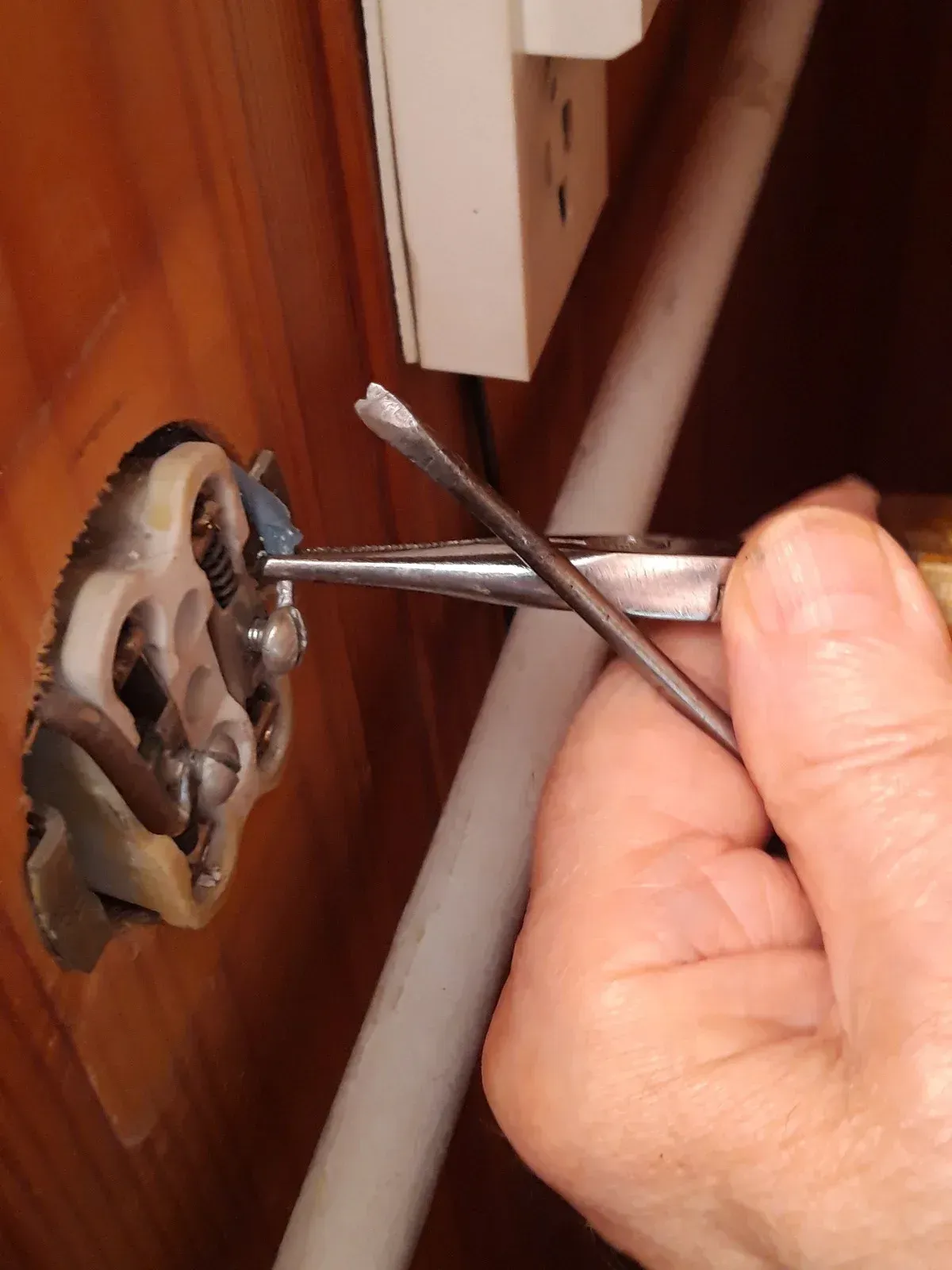 Person using pliers and screwdriver on a dismantled valve, beside a white pipe and electrical outlet.