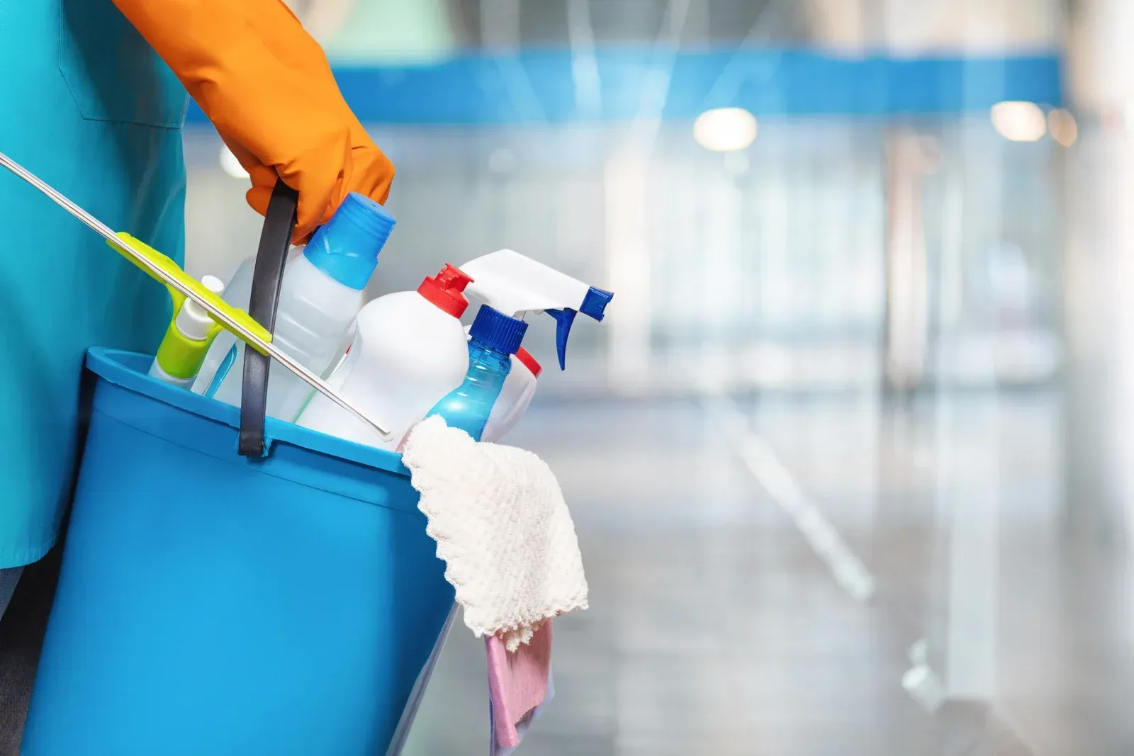Person holding a blue cleaning bucket with various cleaning supplies in a hallway, wearing orange gloves.
