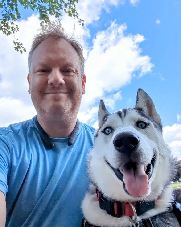 A man in a blue shirt smiling next to a happy, light-colored husky with bright blue eyes against a sunny, blue sky. Dog trainer Northville MI.