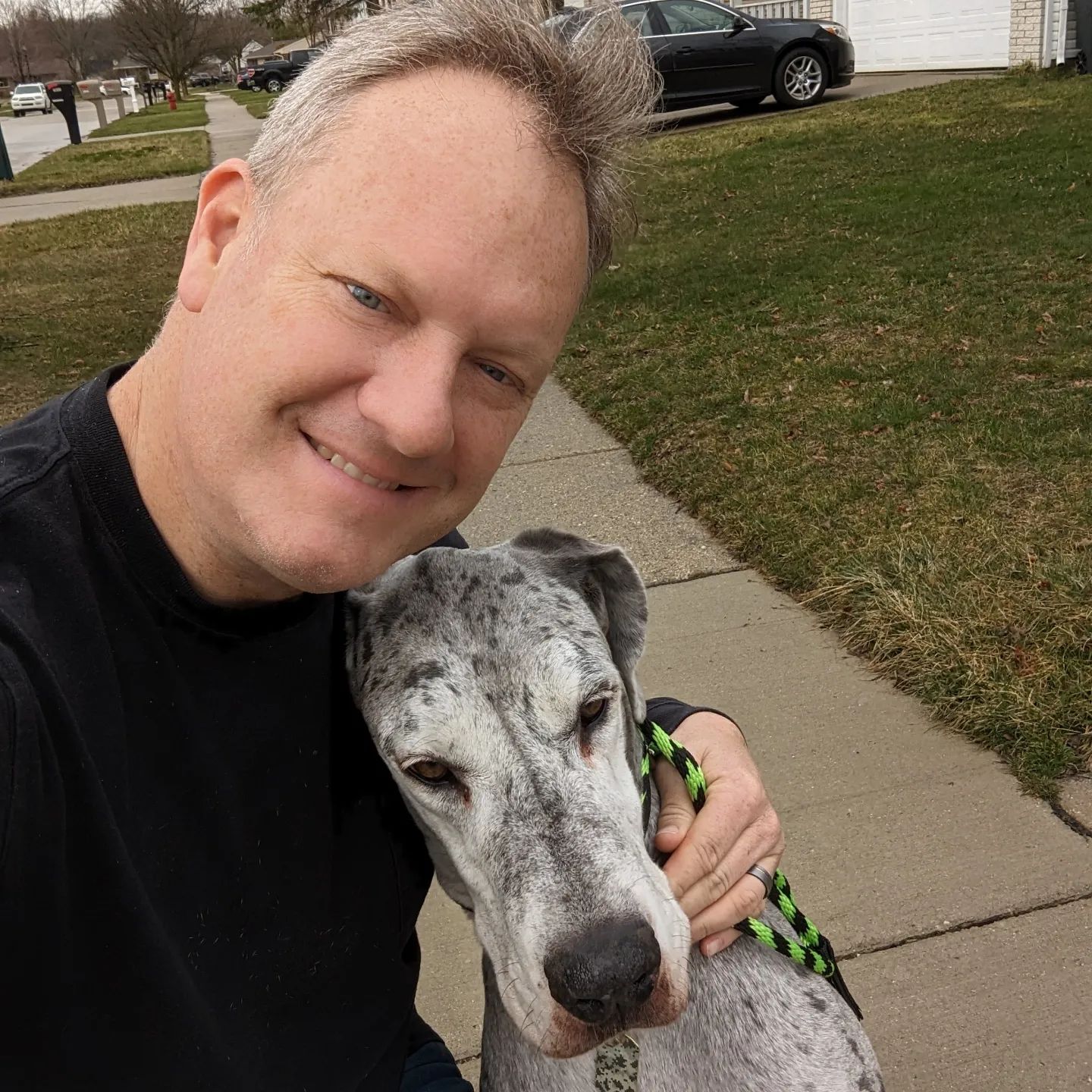 A smiling person hugs a gray and white speckled dog on a sidewalk next to a grassy lawn. Dog trainer in Novi MI.