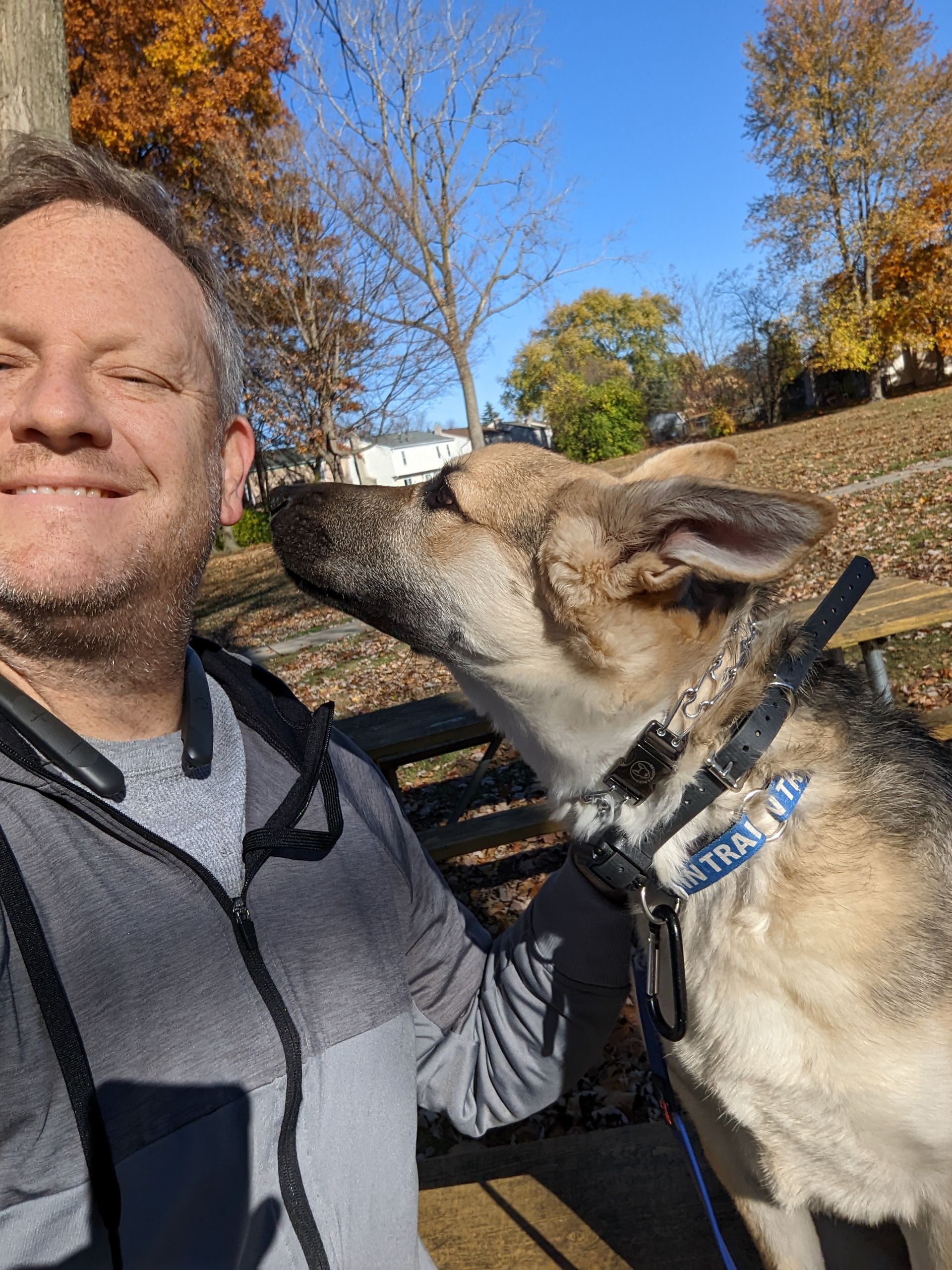 A man smiles for a selfie outdoors in autumn as his dog nuzzles his cheek.